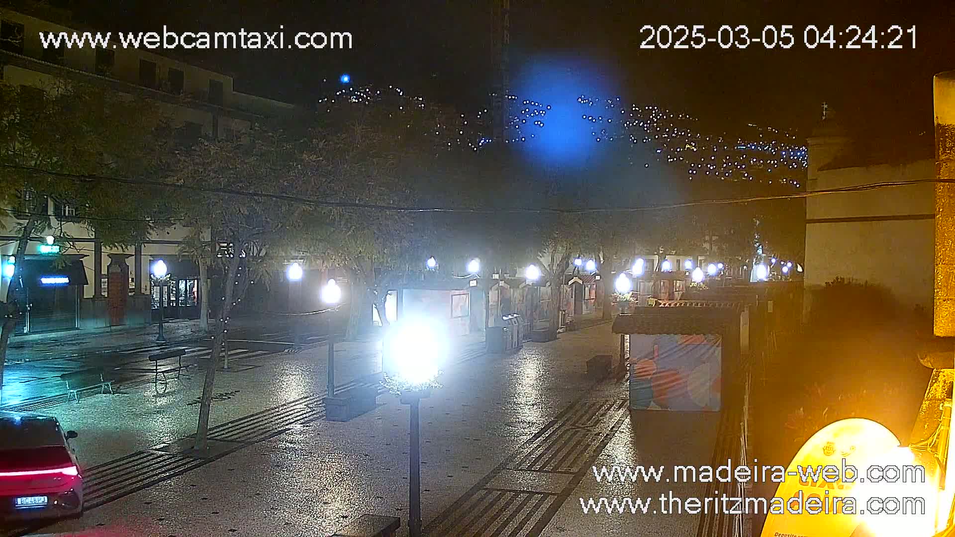 A rain-slicked nighttime plaza, illuminated by streetlights, shows buildings and a few parked cars.