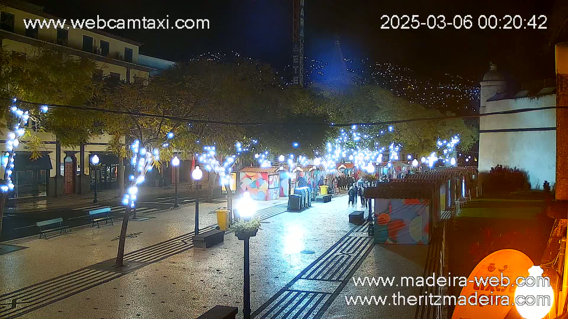 A nighttime view of a town square decorated with string lights, featuring several small market stalls and a few pedestrians.