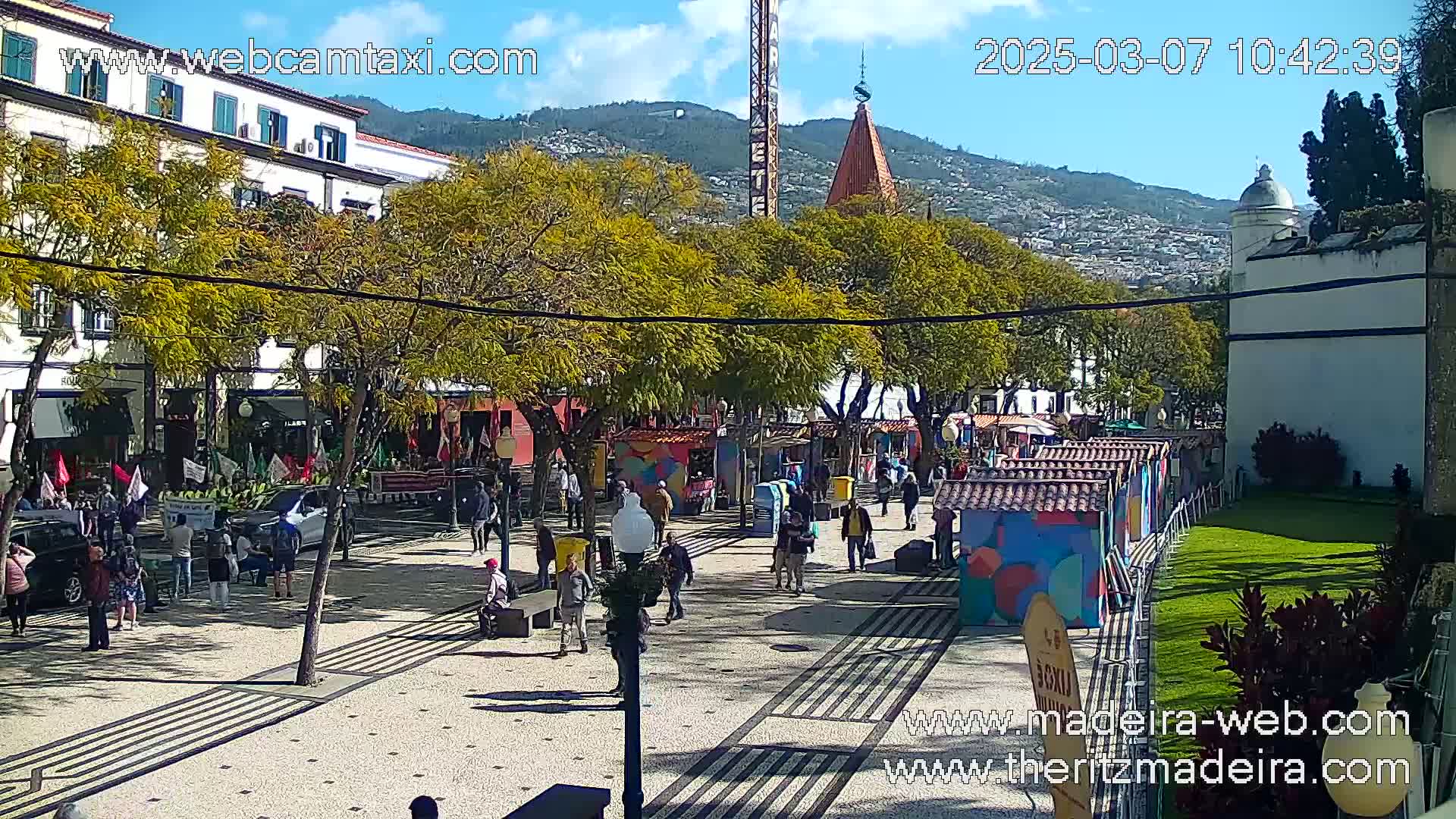 A sunny outdoor plaza scene shows people walking among numerous small colorful stalls and trees, with buildings and mountains in the background.