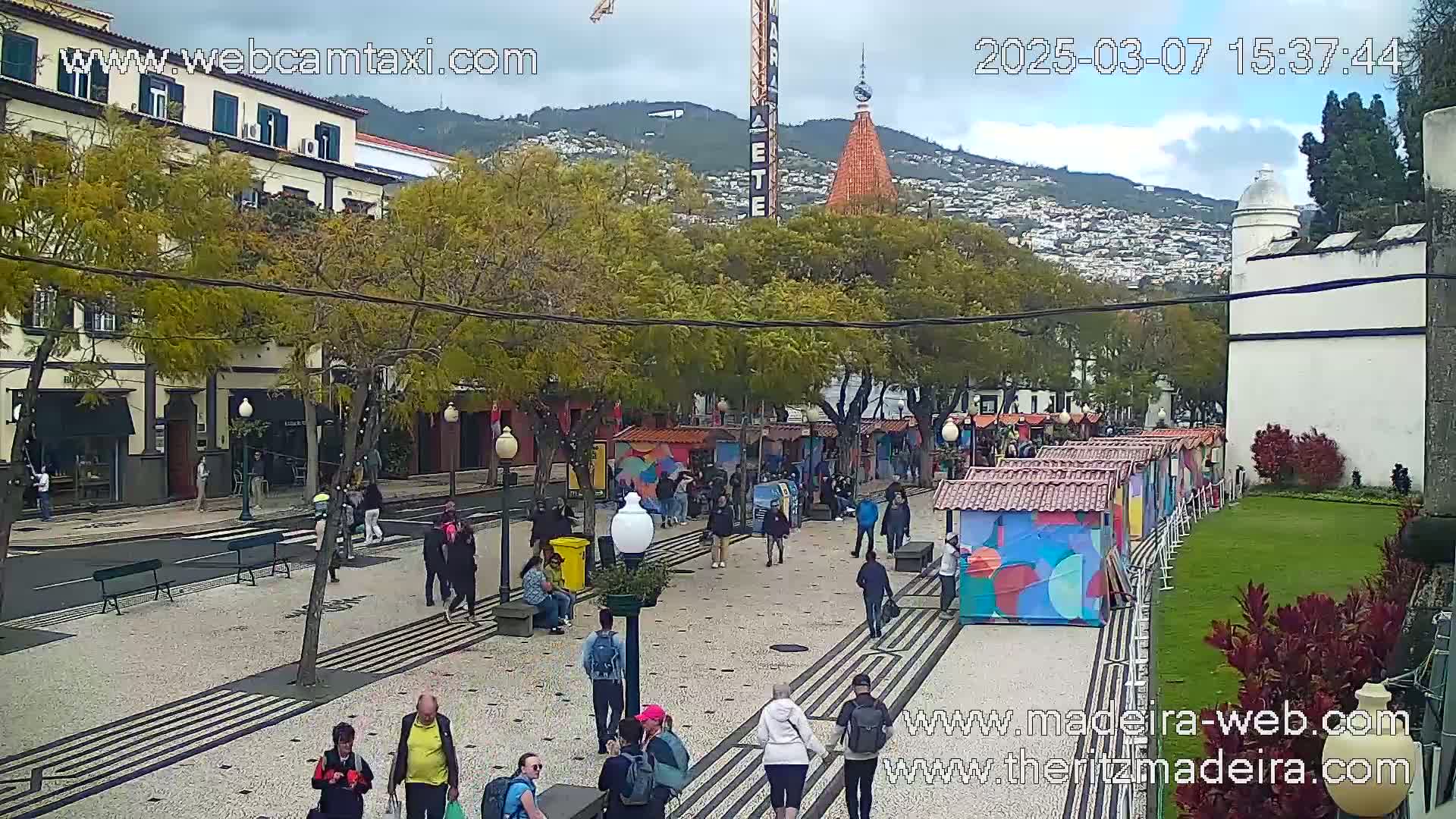 A paved pedestrian area with numerous people walking, lined with colorful market stalls and trees, under an overcast sky.