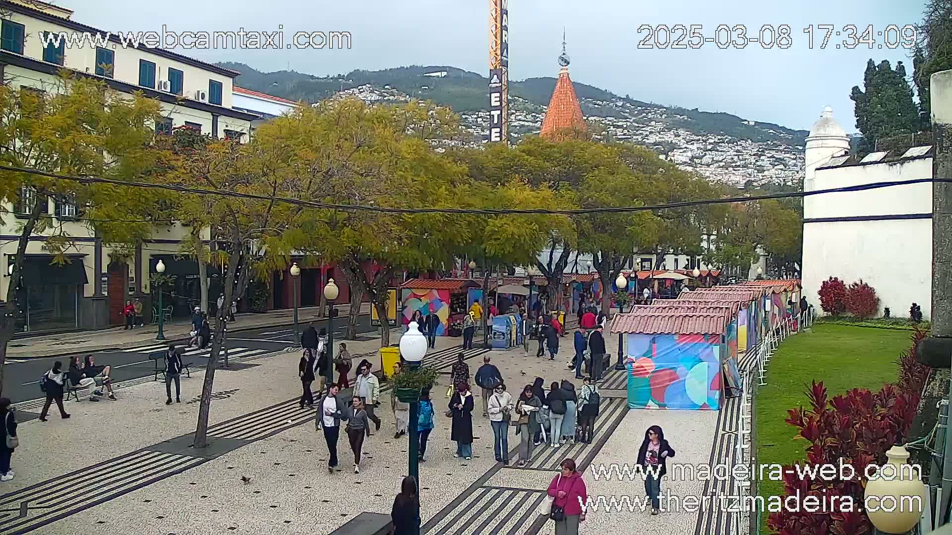 A paved plaza with pedestrians, market stalls, and trees under an overcast sky, with buildings and hills in the background.