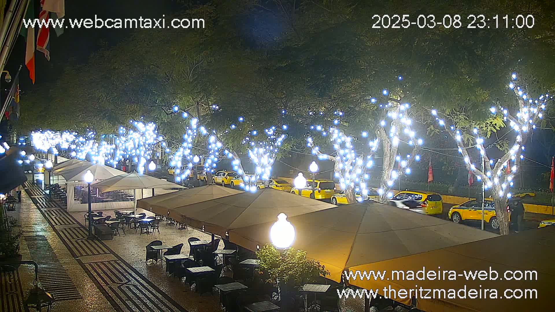 A nighttime outdoor scene shows a paved pedestrian area with cafe umbrellas and tables under trees strung with white lights, and yellow taxis parked along the street.