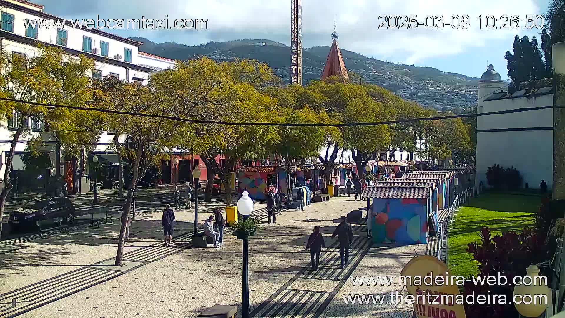 A sunny outdoor plaza scene shows several people walking among colorful market stalls and trees, with mountains in the background.