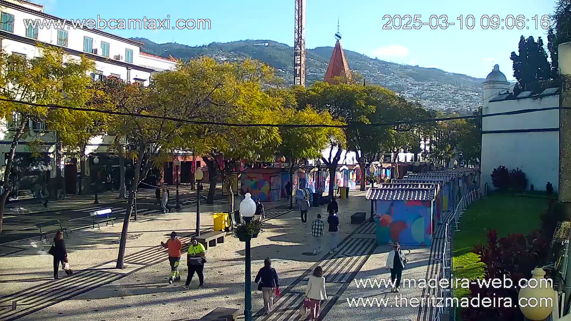 A paved plaza with several people walking, surrounded by trees, small colorful stalls, and buildings, under a sunny sky with mountains in the background.