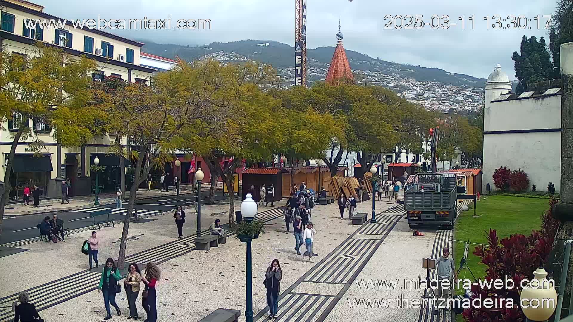 A paved plaza with people walking, trees, buildings, and a flatbed truck, under a partly cloudy sky.
