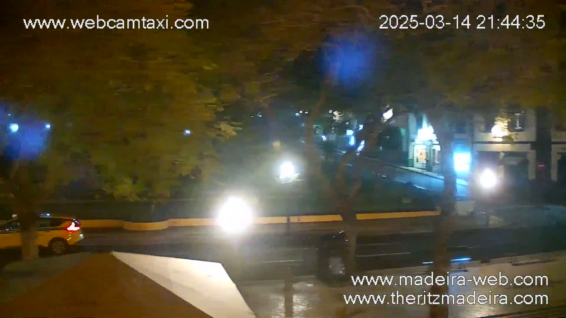 A nighttime street scene shows a yellow car and other vehicles on a wet road, passing buildings with lights on under leafy trees.