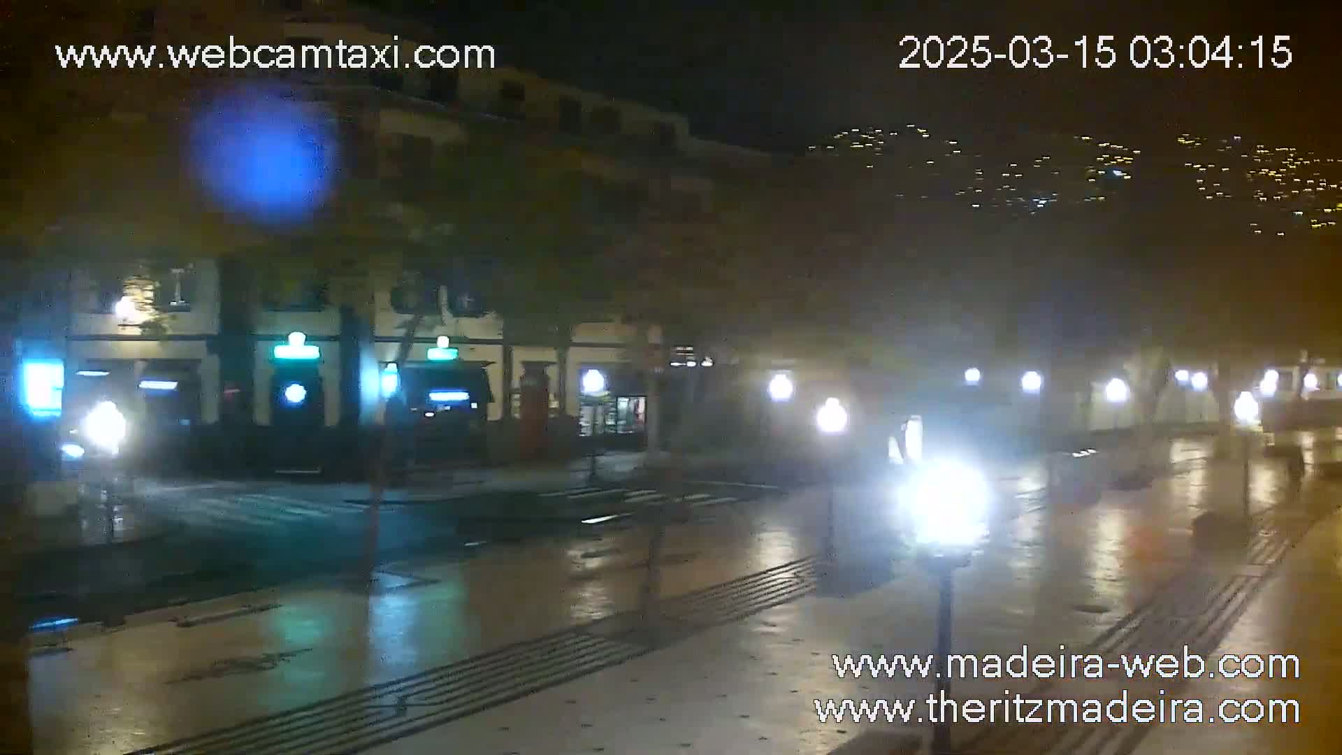 A wet, nighttime street scene shows a row of buildings with illuminated signs, streetlights reflecting on the pavement, and distant lights on a hillside.