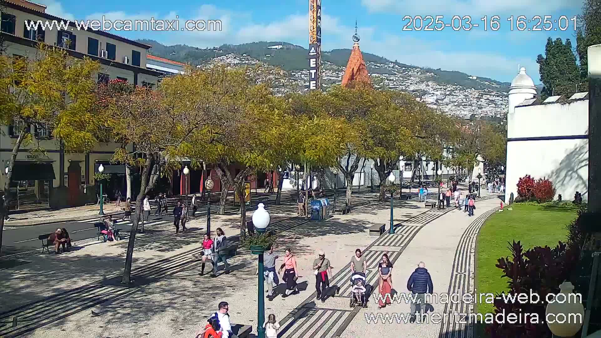 A sunny day reveals a pedestrian plaza with people walking along a patterned walkway, lined with trees and buildings, and hills visible in the background.