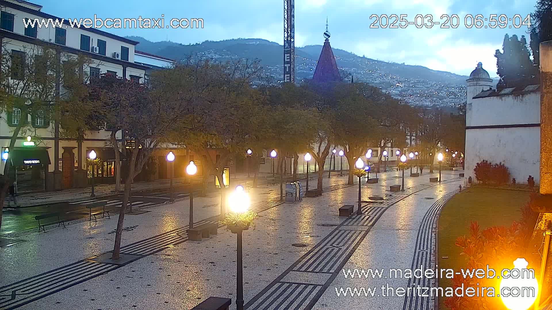 A wet, paved plaza at twilight is illuminated by streetlights, lined with trees and buildings, with a hill visible in the background.