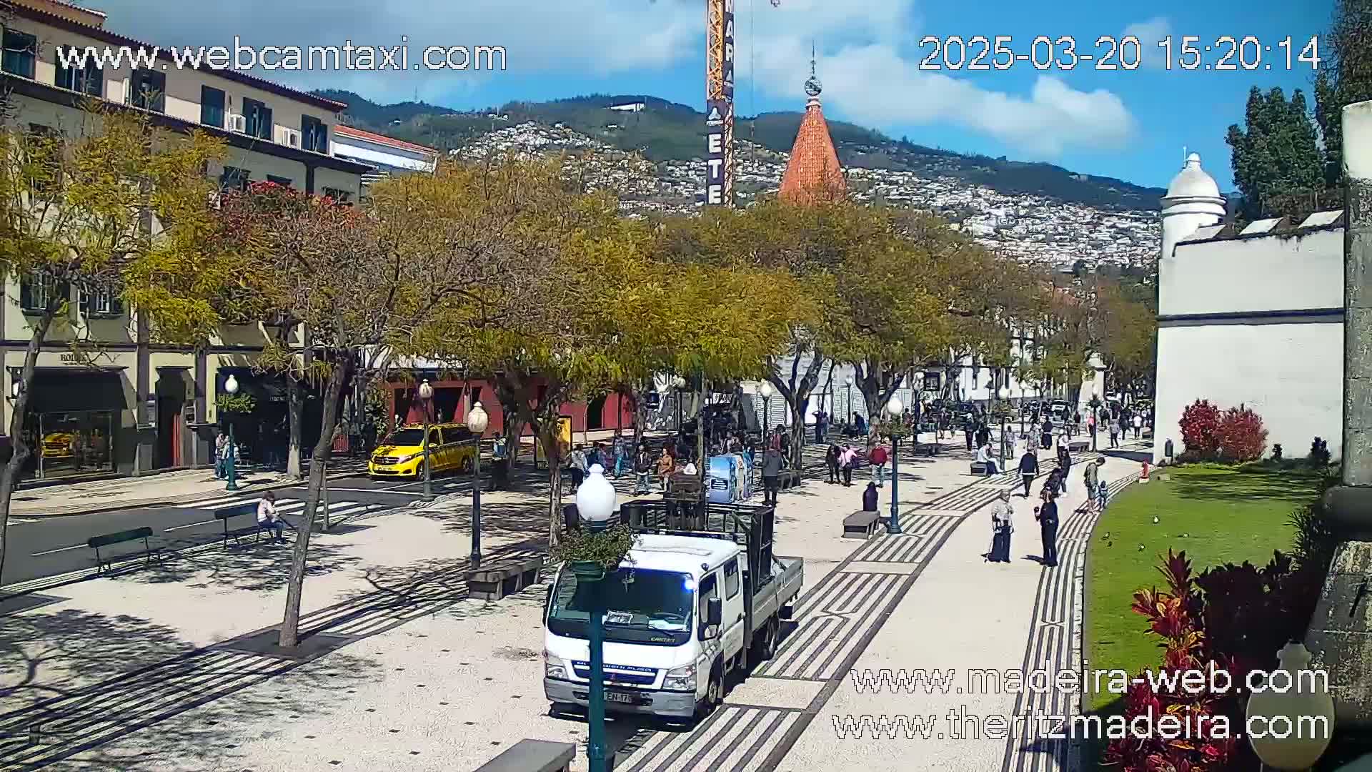 A sunny city square with pedestrians, parked cars, and a white pickup truck, bordered by buildings and trees.