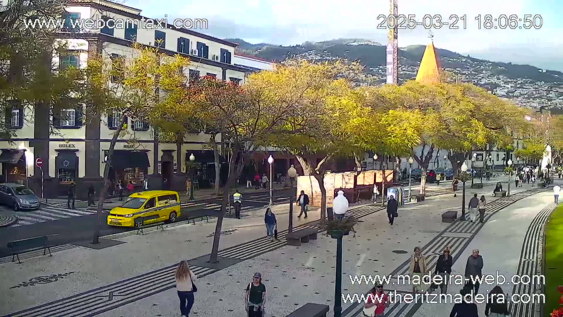A paved pedestrian area with people walking, lined with trees and buildings, under a mostly clear sky.