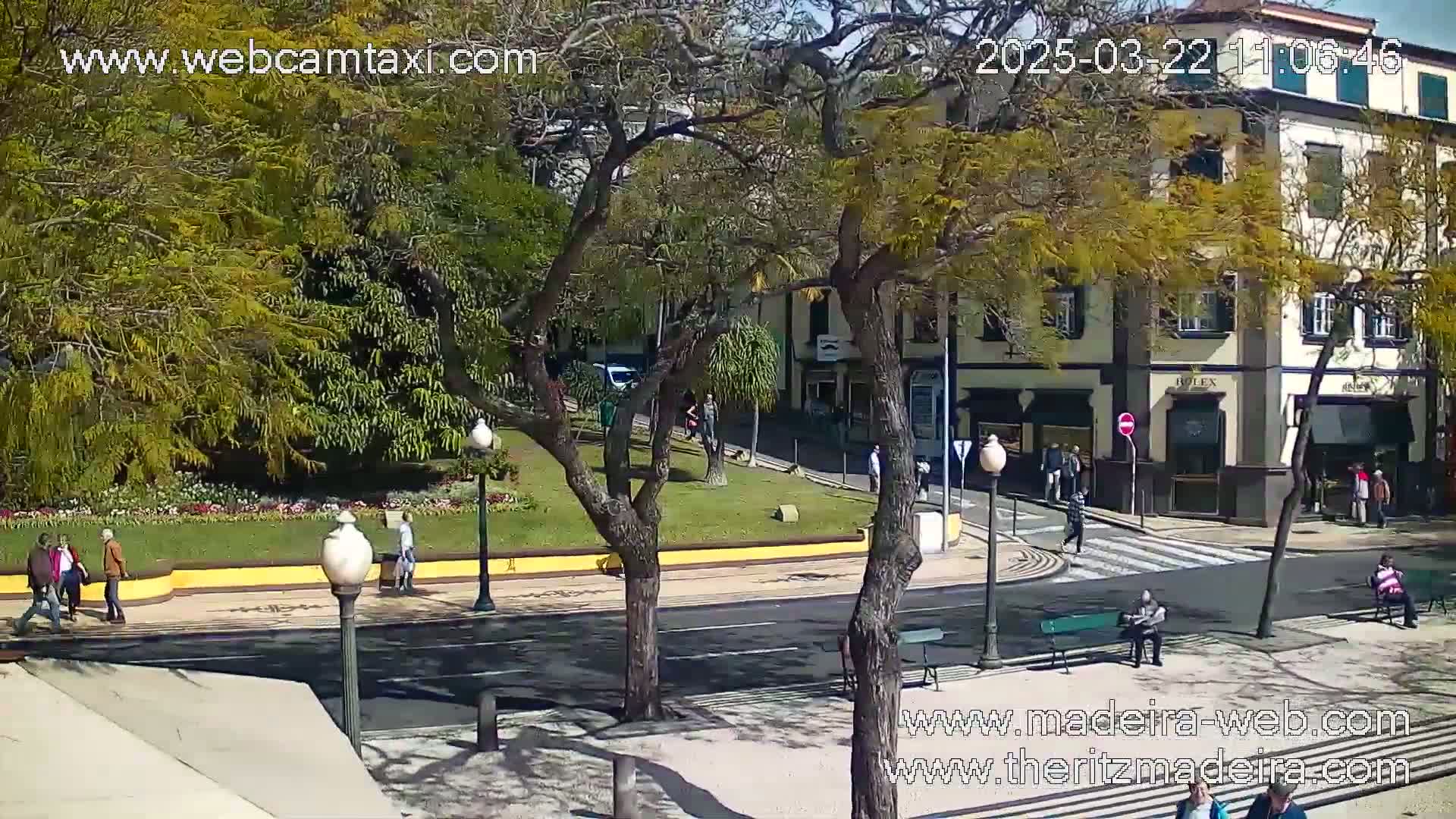 A sunny street scene shows a paved road with pedestrians, a small park with trees and flowers, and a multi-story building with shops on the ground floor.