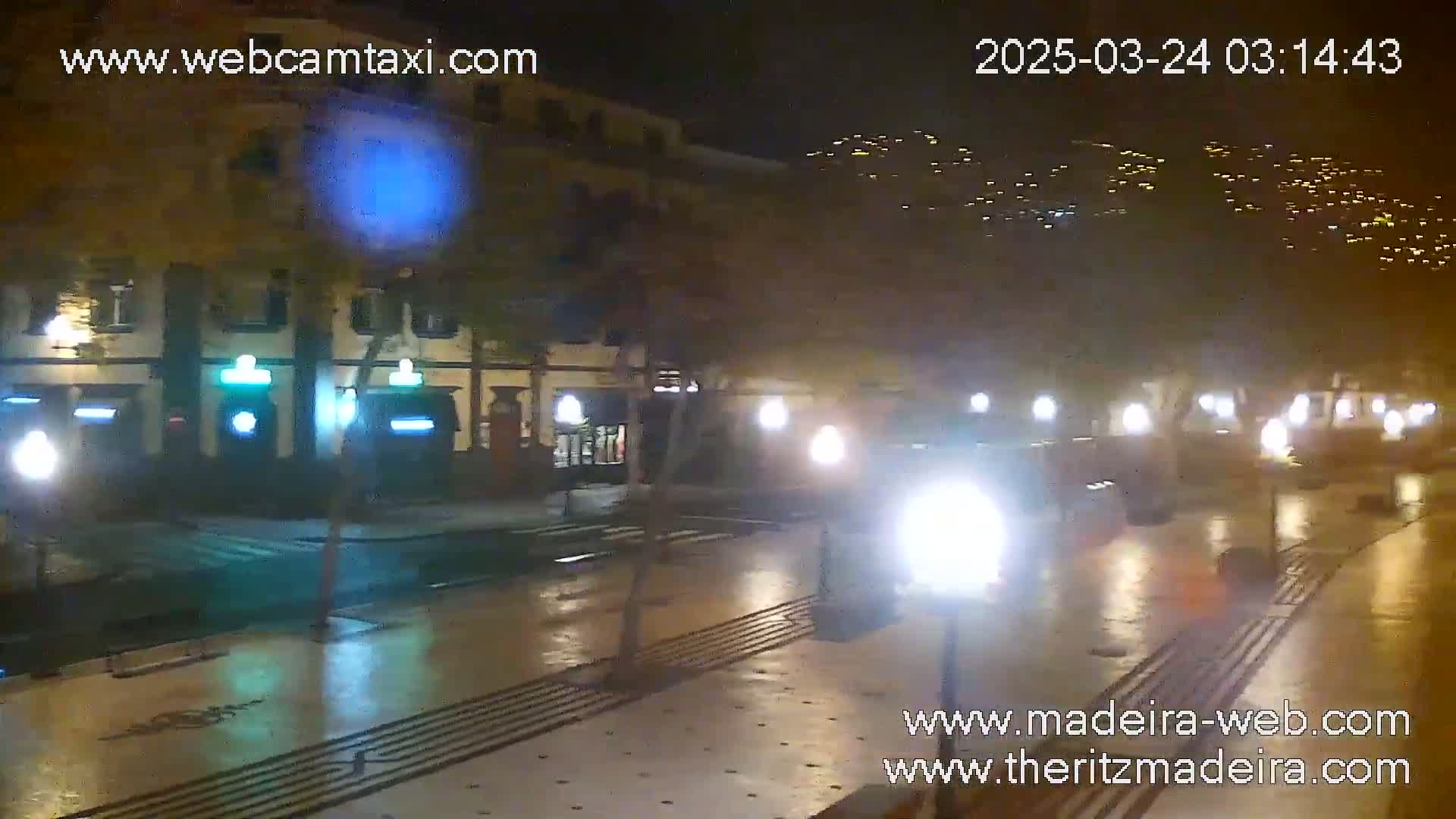 A wet, nighttime street scene shows a building with lights on, several cars driving down the street, and streetlights illuminating a paved area.