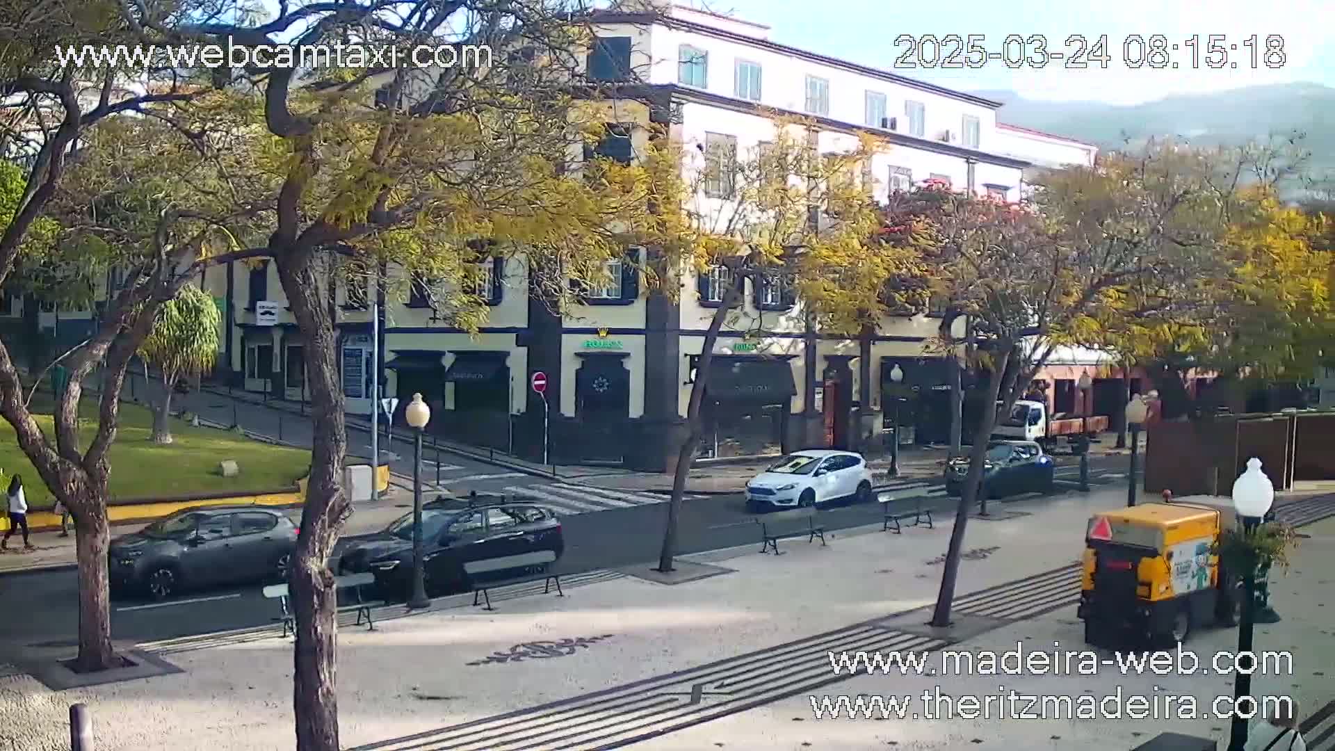 A street scene on a sunny day shows several cars parked and driving near a multi-story building, with large trees and a street cleaning vehicle visible.