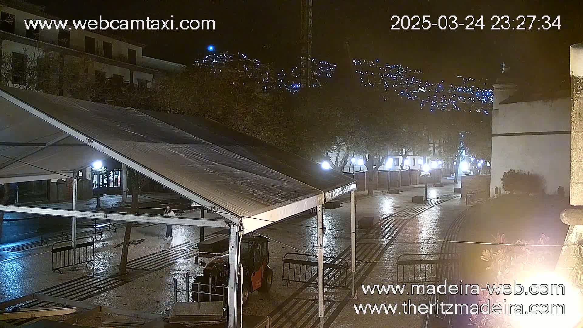 A wet, nighttime town square is seen under a partially covered tent, with a hill of lights visible in the background.