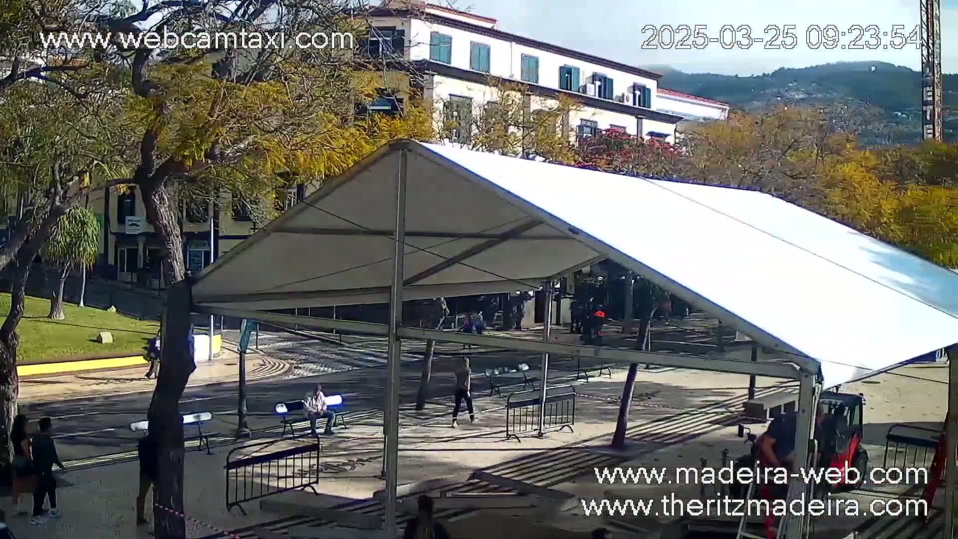 A white tent structure stands in a paved area, near a street and buildings, under a sunny sky.