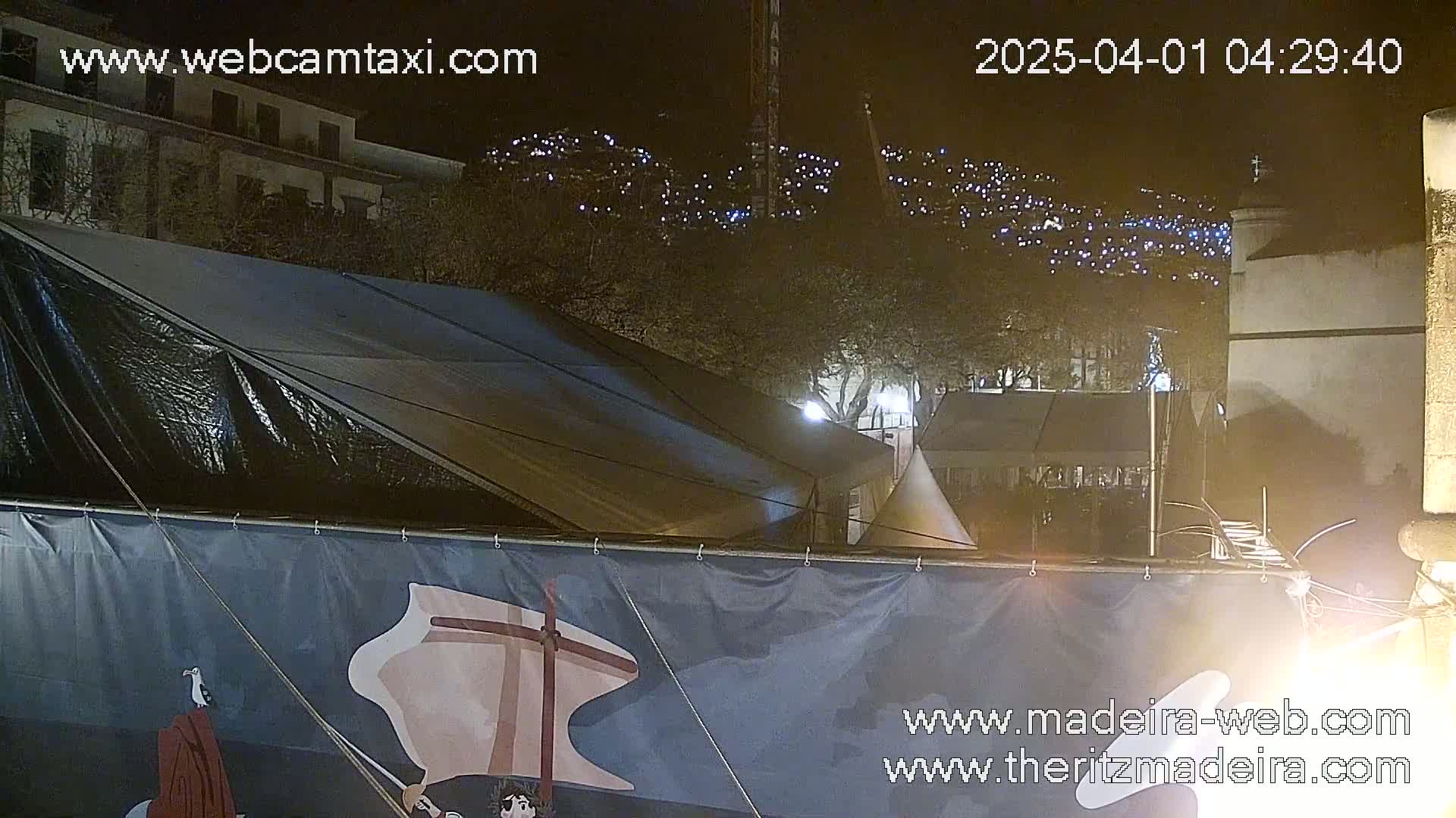 A nighttime view of a partly covered tent with a large banner in front, overlooking a hillside illuminated with numerous small lights.