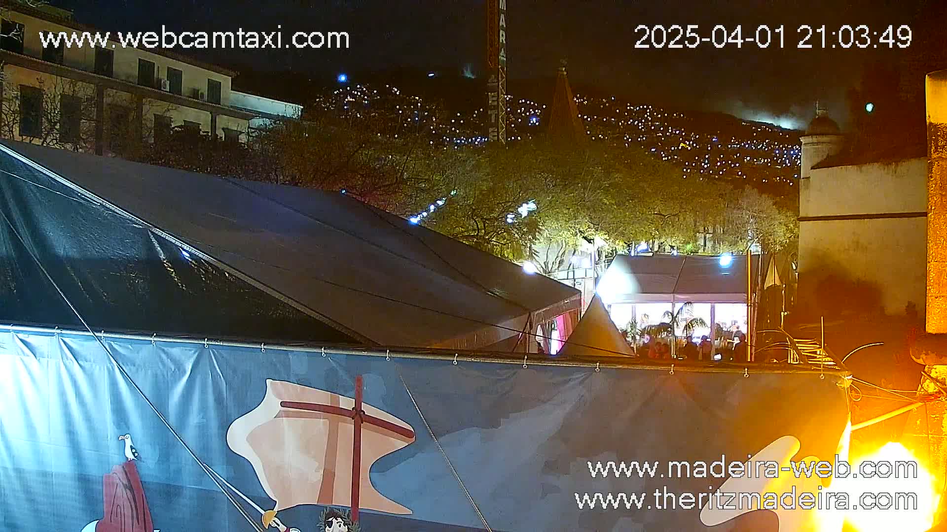 A nighttime view of a large tent with a painted banner in front, partially obscuring a view of a hill covered in lights and buildings beyond.