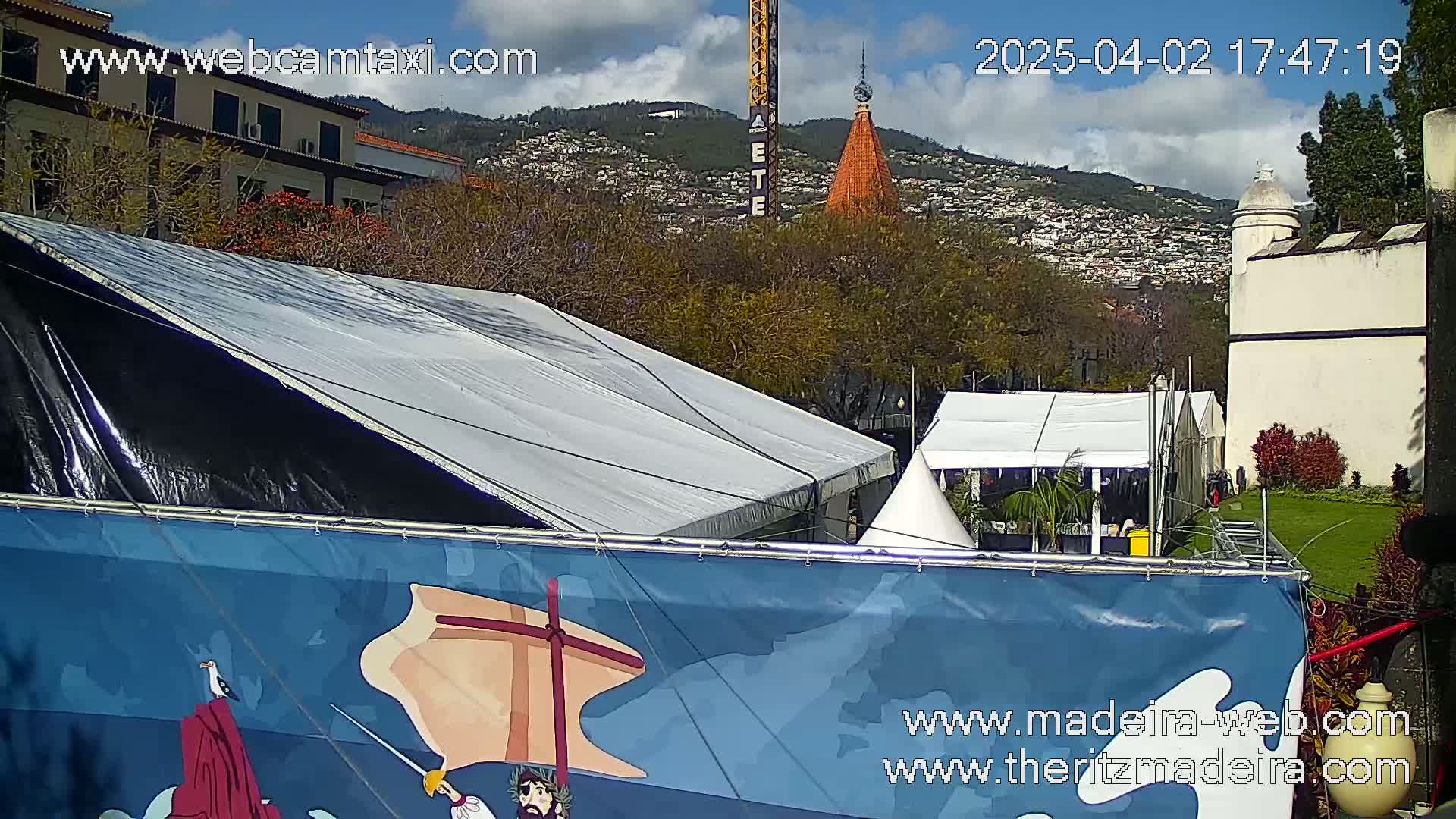 A large tent with a blue banner depicting a ship and figures stands before a white building under a partly cloudy sky.