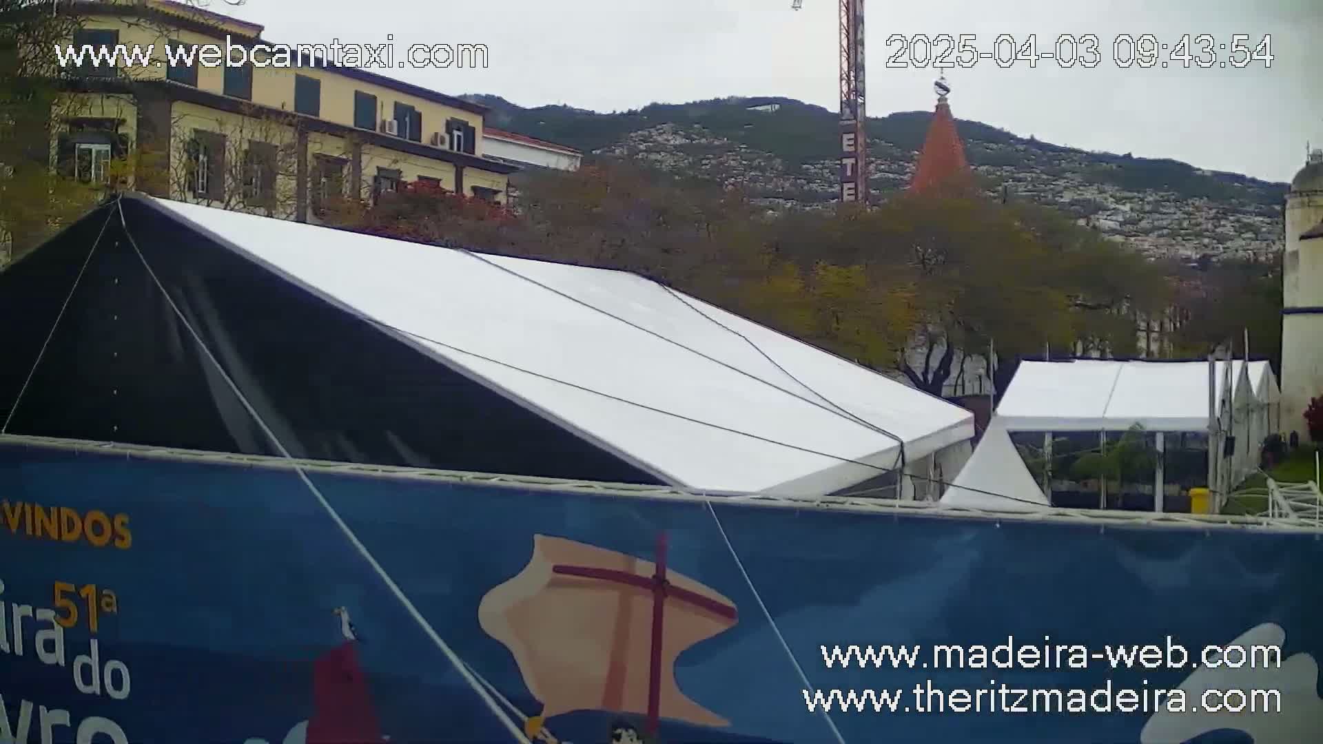 A large white and black tent is set up in front of a blue banner, with other smaller white tents visible behind it and buildings and hills in the background under an overcast sky.