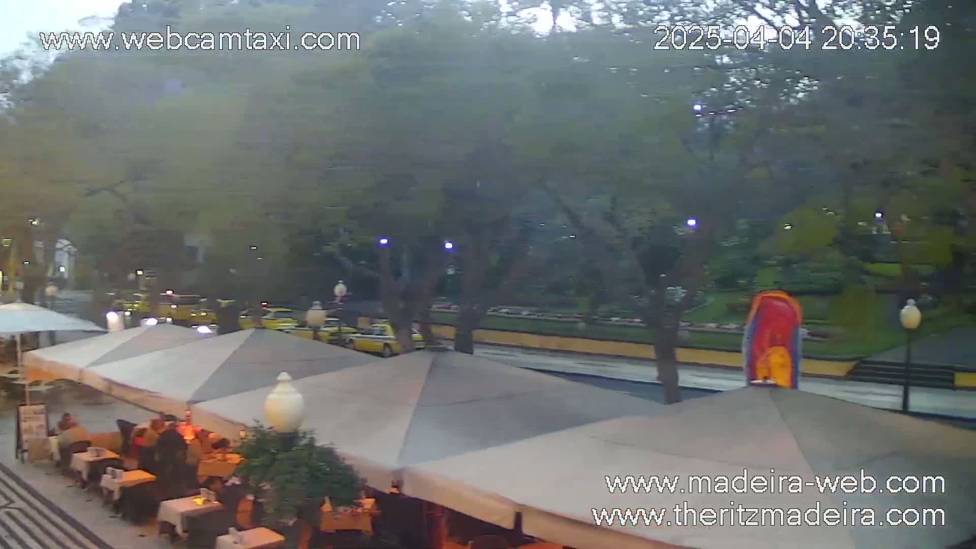 An outdoor cafe with several umbrellas and patrons sits beside a street with cars driving past under overcast skies.