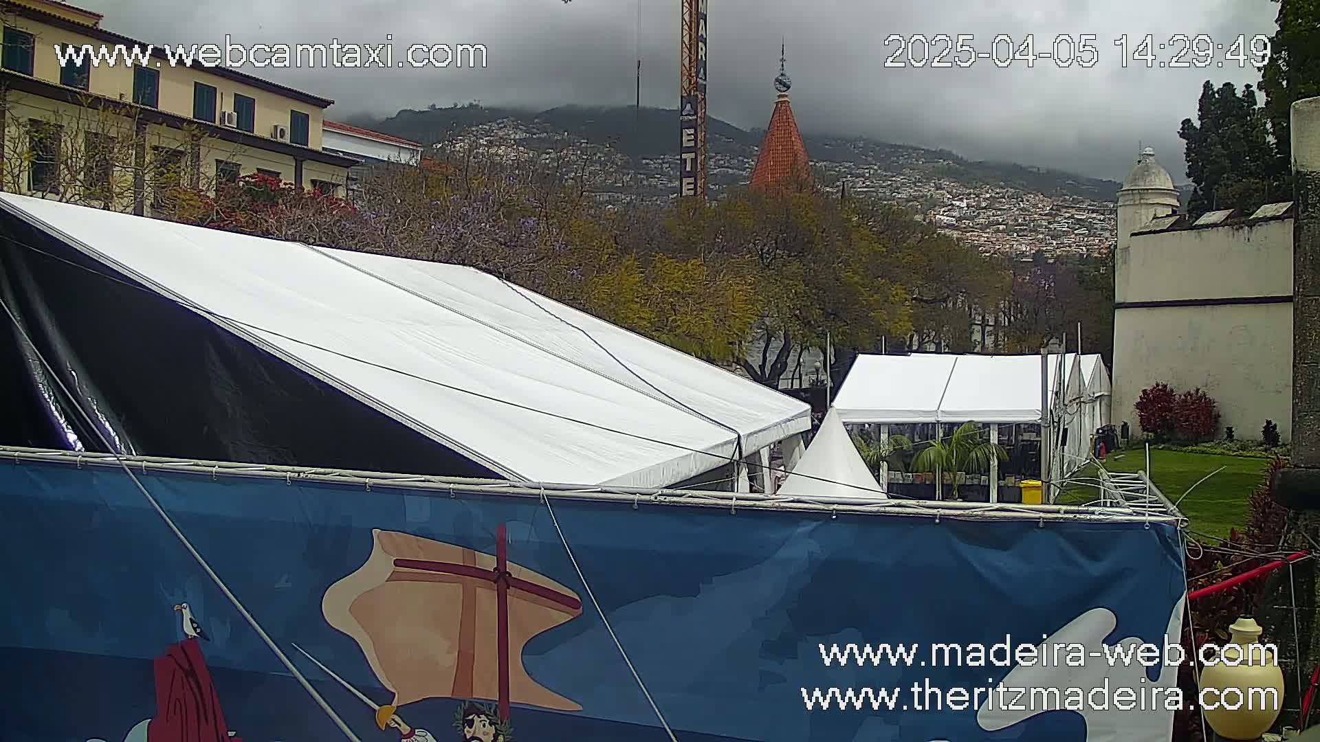 A large white and black tent is situated in front of a blue banner, with other smaller tents visible in the background under an overcast sky; a town and mountains are in the distance.