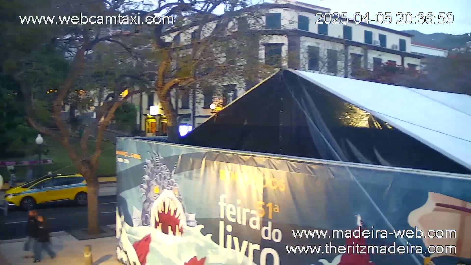 A yellow taxi is parked on a city street next to a large tent-like structure with a banner, under a twilight sky.