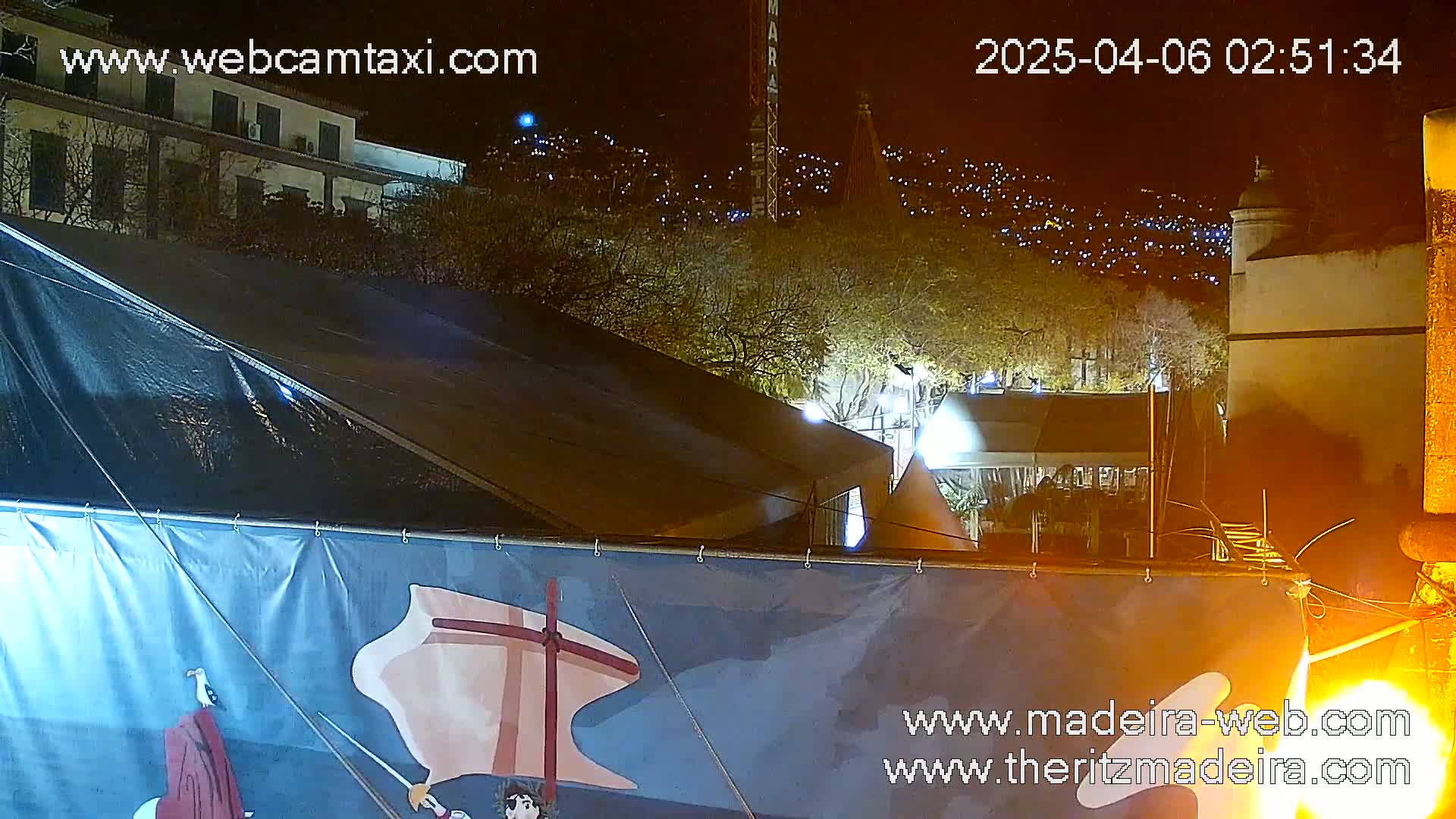A nighttime view shows a dark-colored tent with a painted banner in front of it, situated in front of a hill dotted with lights, with a building and trees visible in the background.