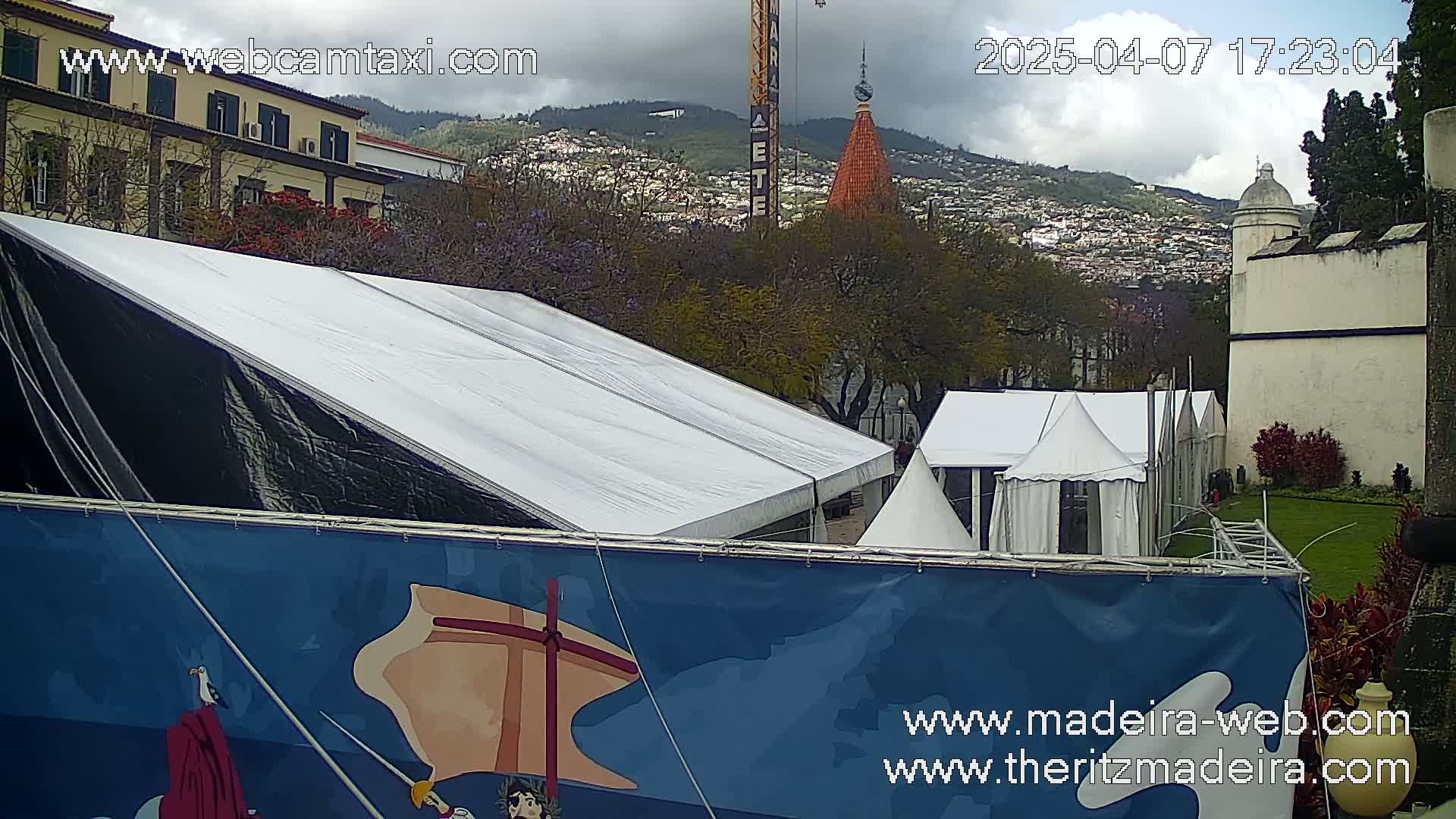 A large white and black tent stands in front of a blue banner depicting a sailing ship, with several smaller white tents behind it, all situated on a grassy area overlooking a town nestled in a cloudy mountain range.