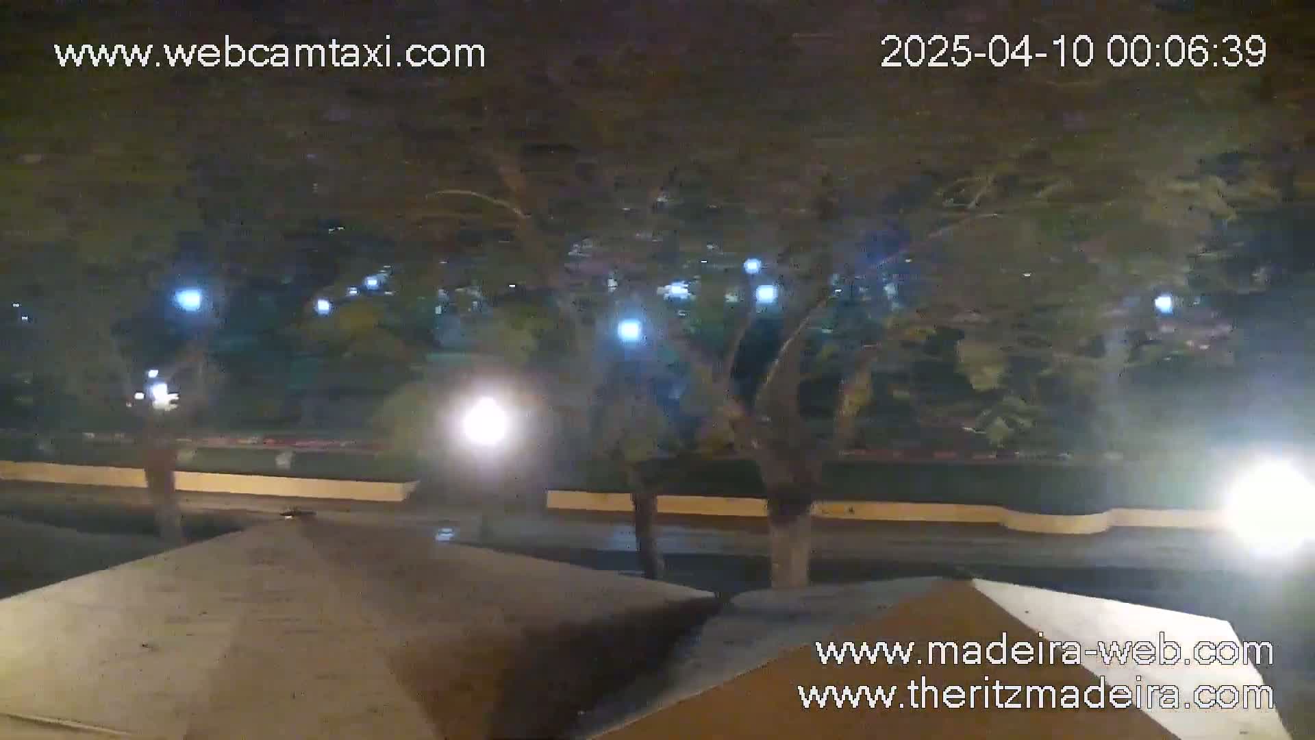 A nighttime, outdoor scene shows a street with trees, lights, and a low wall, partially obscured by the angle of what appears to be a security camera.