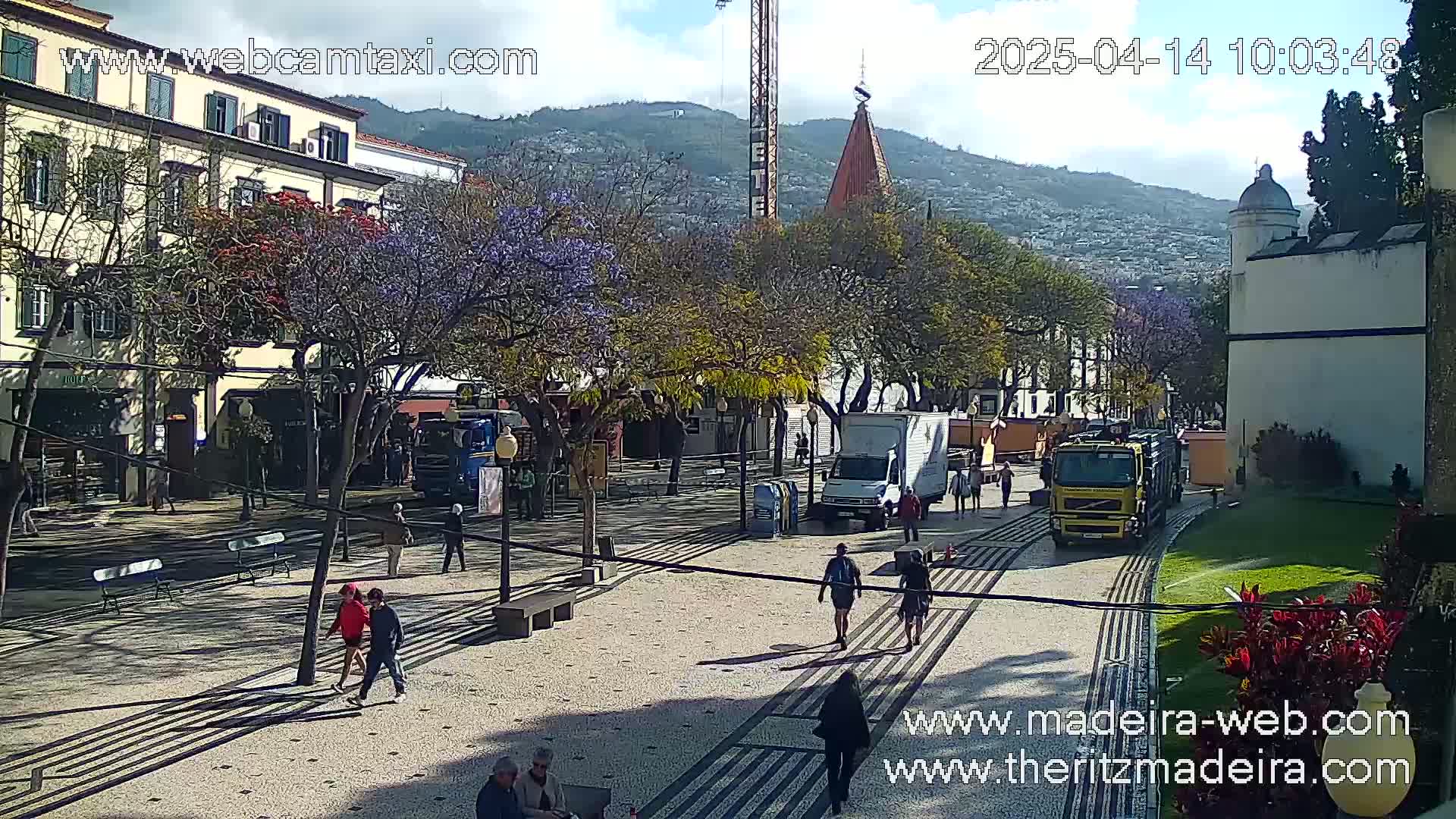 A sunny plaza scene shows people walking among trees and buildings, with a few delivery trucks parked nearby, and a mountain in the background.