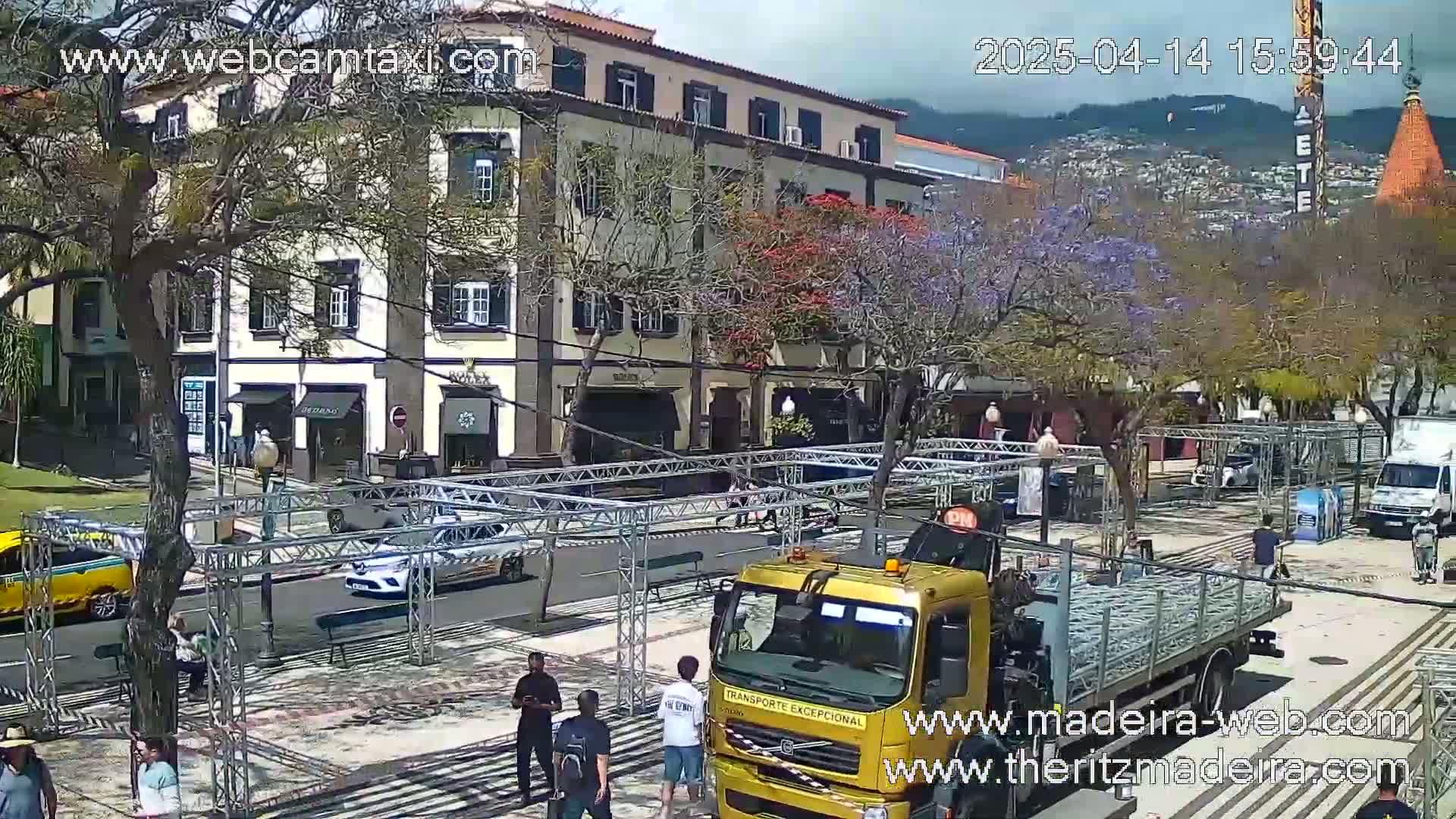 A sunny day shows a town square with a large metal structure being assembled, a gold truck carrying more metal, and several people milling about near multi-story buildings and trees with purple blossoms.