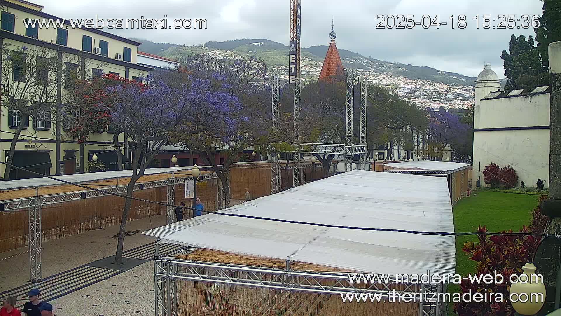 An overcast day shows a town square with several market stalls under temporary roofs, flowering trees, and buildings, with hills in the background.