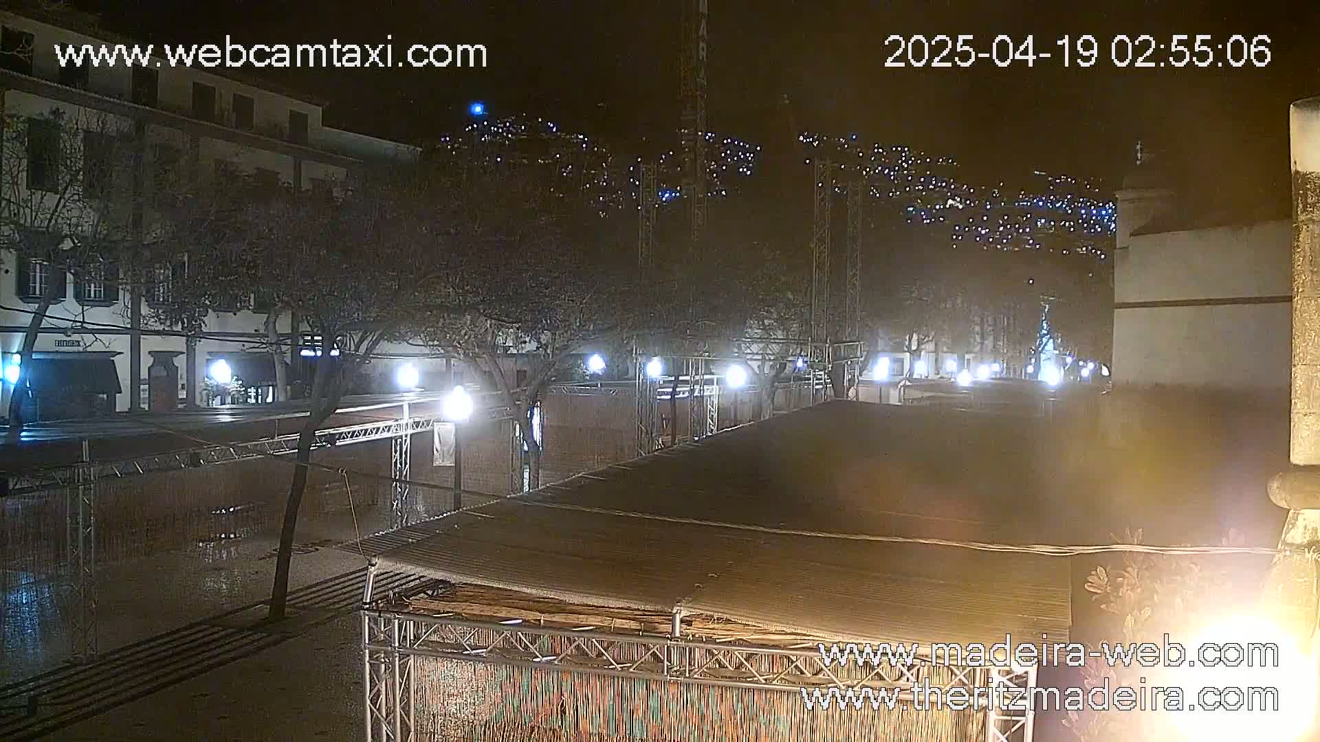 A nighttime view of a town square with several buildings, trees, and temporary structures, illuminated by streetlights and other lights on buildings in the background.