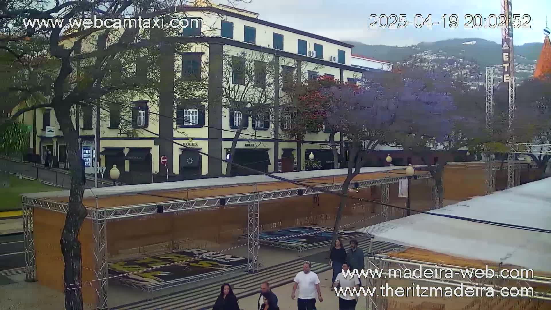 A multi-story building with several businesses on the ground floor is visible under an overcast sky, with a street scene including several people and temporary structures in the foreground.