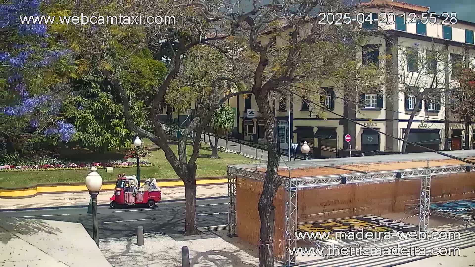A red three-wheeled vehicle drives on a street past a park with flowering trees, next to a building, under a partly cloudy sky.
