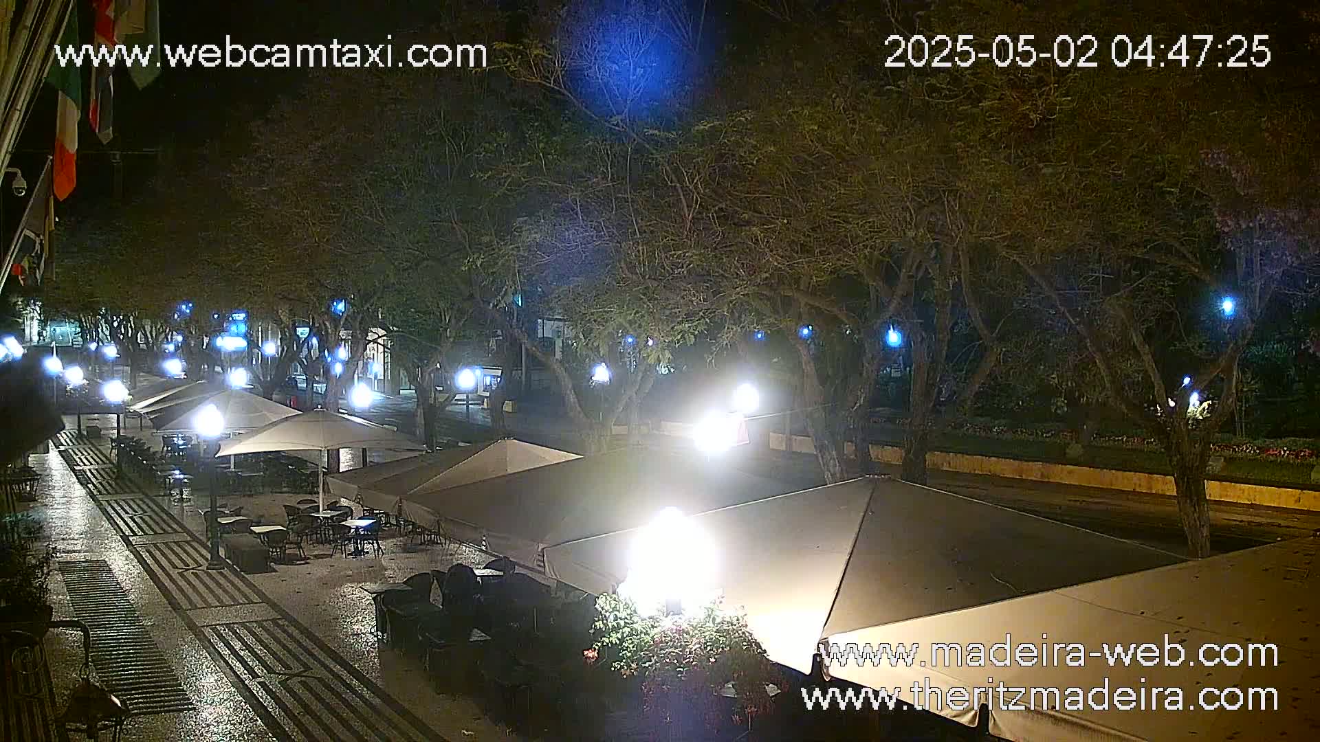 A nighttime outdoor scene shows a wet, paved plaza with several cafe umbrellas and tables under illuminated trees.