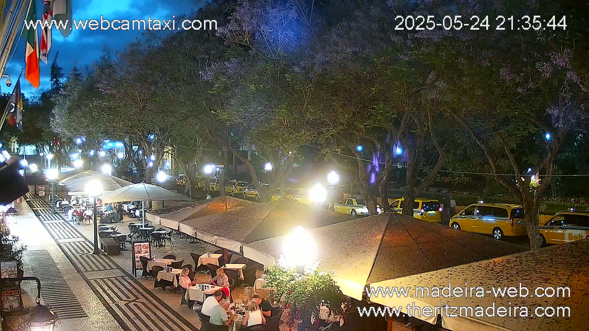 An outdoor nighttime scene shows a paved pedestrian area with outdoor seating at restaurants under umbrellas, lined with trees strung with lights, and yellow taxis driving along a road.