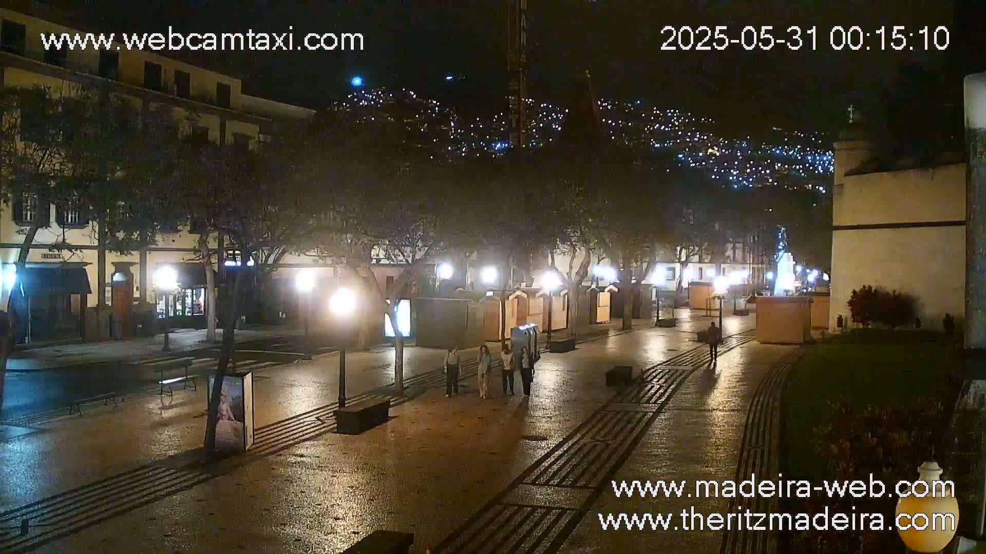 A wet, nighttime street scene shows a paved plaza with people walking, illuminated by streetlights and the lights of buildings, with a hillside of lights in the background.