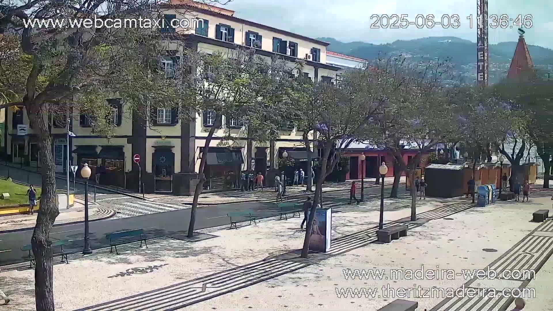 A sunny day reveals a paved plaza with benches and several people standing near a multi-story building and trees, all under a mostly clear sky.