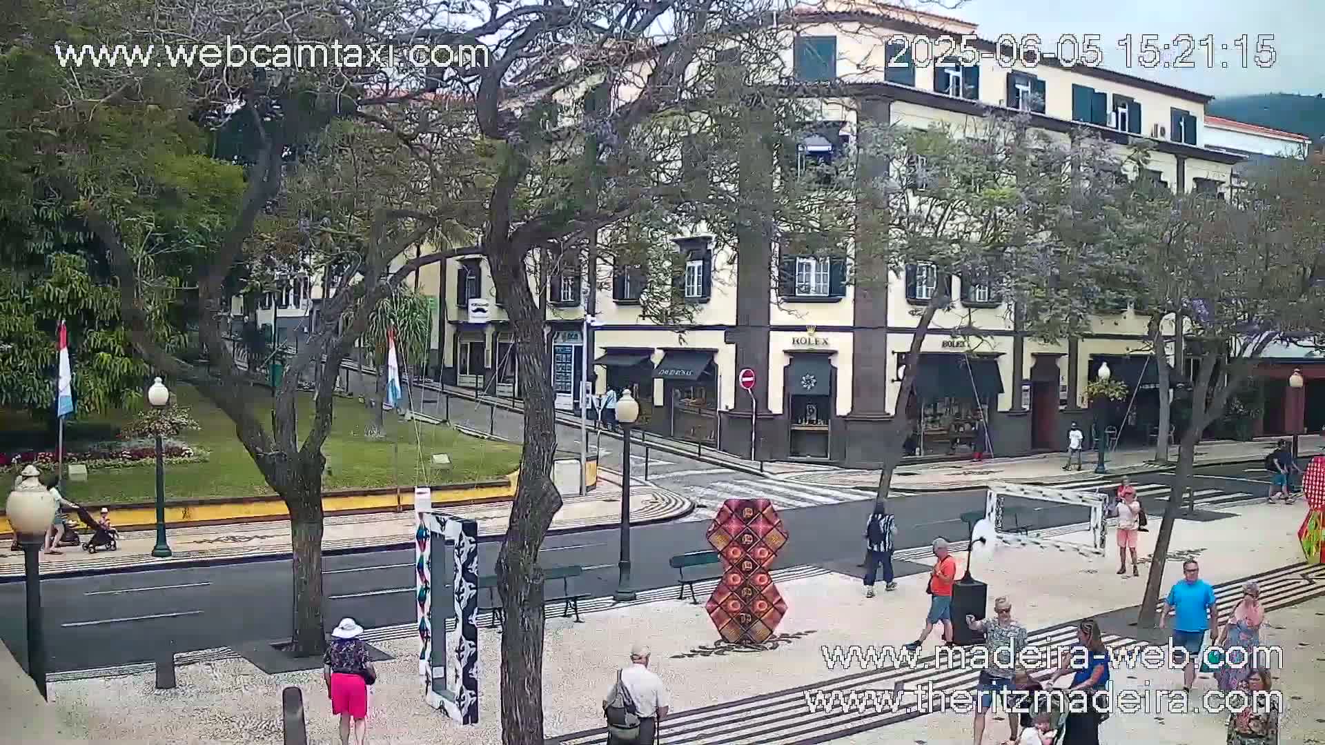 A partly sunny street scene shows a pedestrian area with several people walking past shops and a small park.