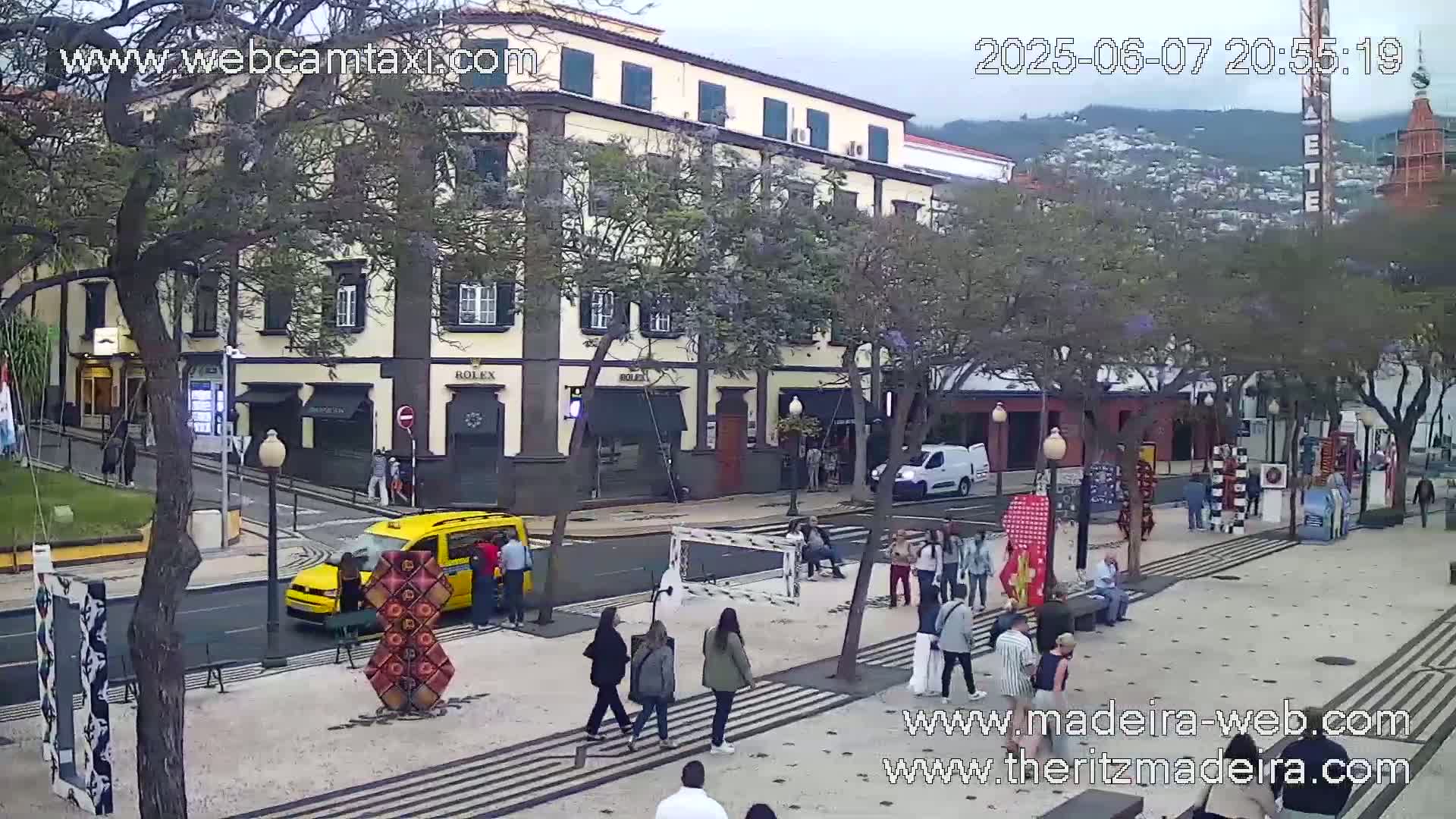 A street scene shows a multi-story building, people walking on a sidewalk, a yellow taxi, and several trees under an overcast sky.