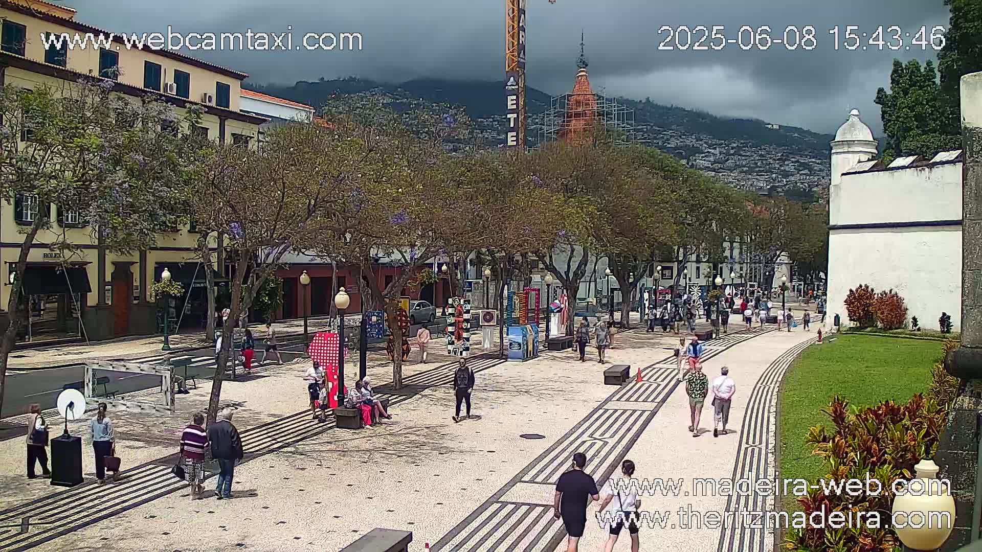 A paved pedestrian area with people walking, lined with trees and buildings, under an overcast sky.