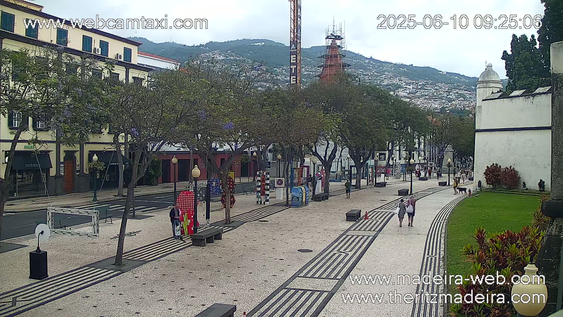 A paved pedestrian walkway lined with trees and art installations stretches towards a hilltop town under an overcast sky.