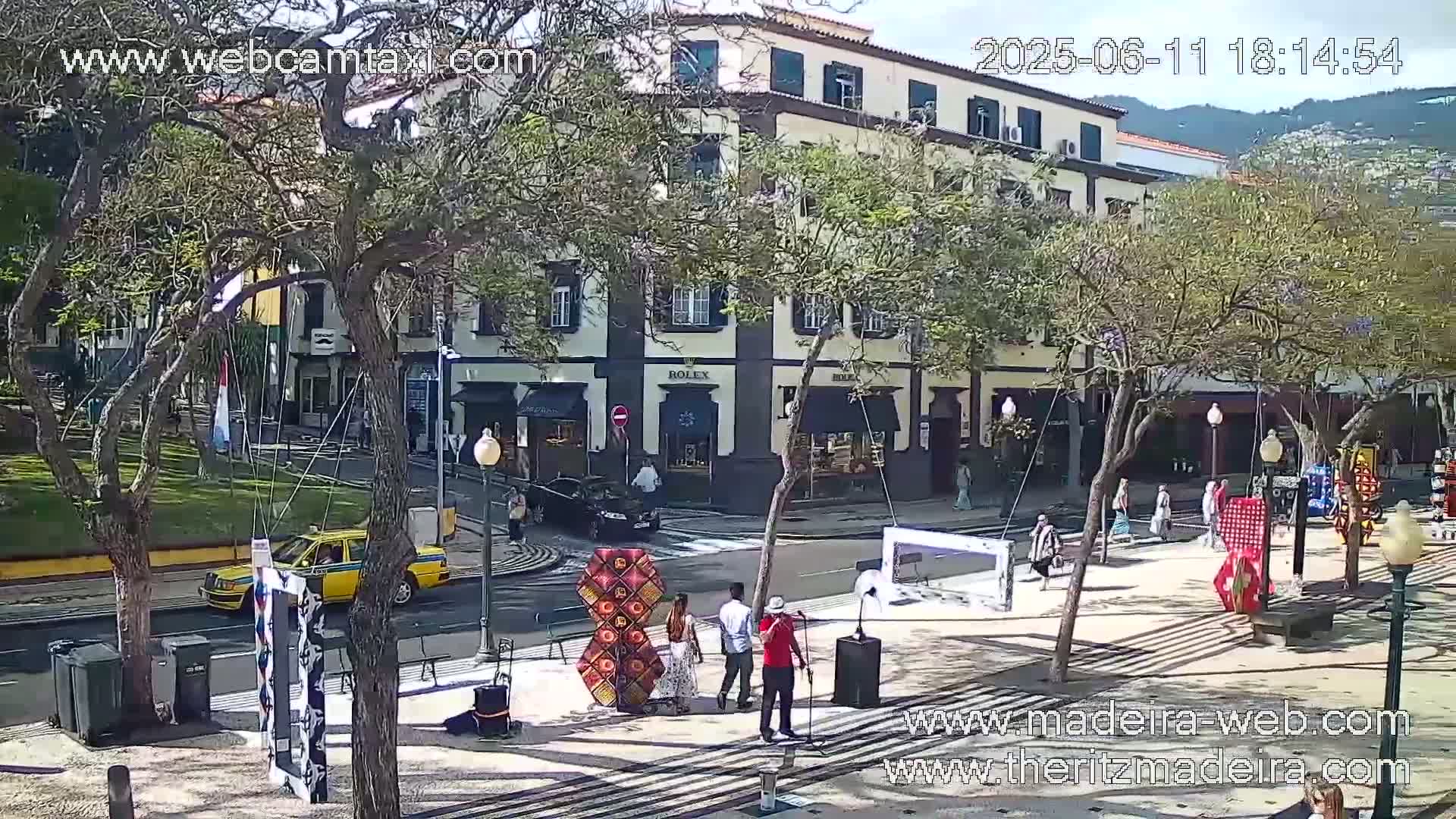 A sunny city square scene shows pedestrians walking past shops and a yellow taxi, with some art installations and a small group of people standing near a microphone setup.