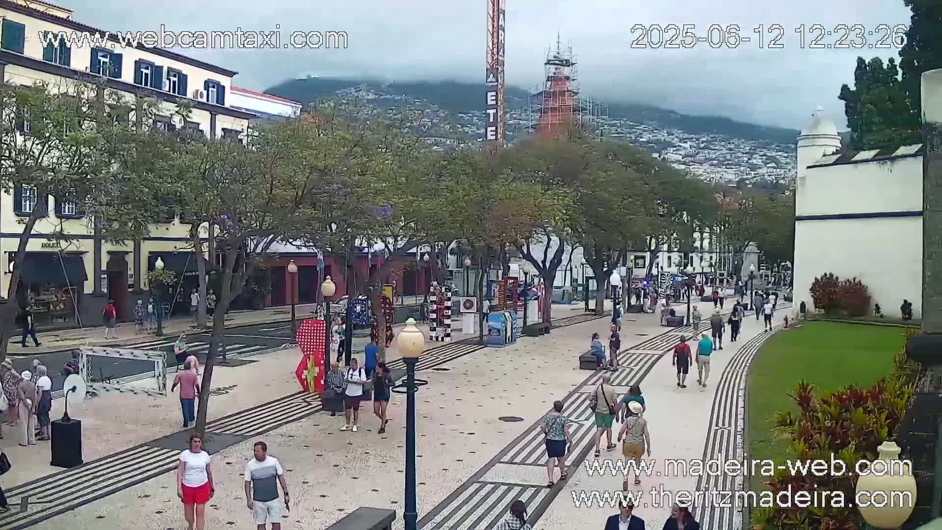A paved pedestrian area with people walking, lined with trees and buildings, under an overcast sky.