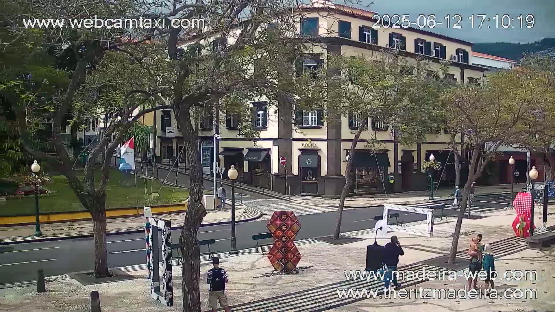 A partly cloudy day shows a street scene with a multi-story building, several pedestrians, and various art installations in a plaza.
