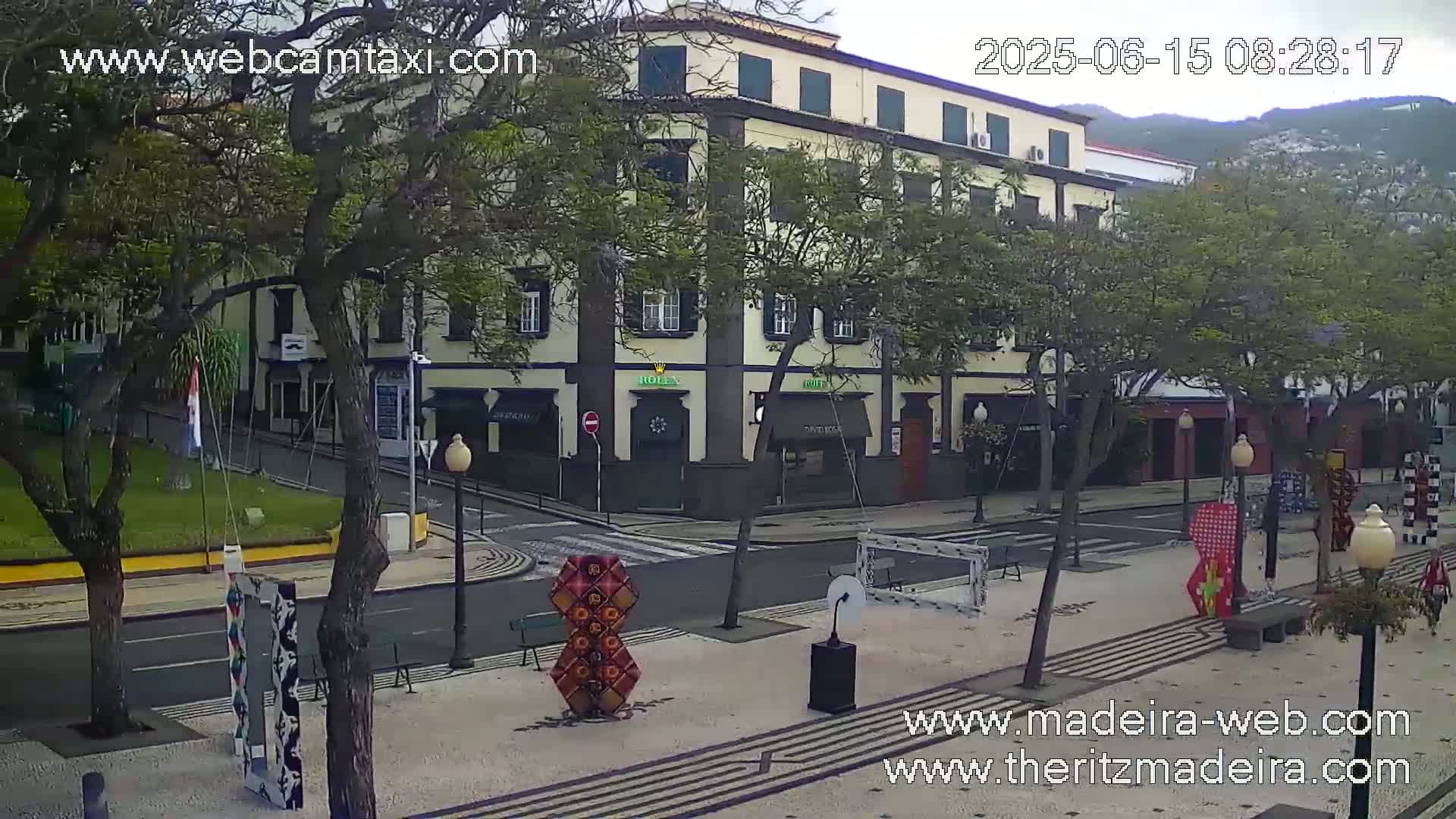 A partly cloudy day shows a town square with a multi-story building, several trees, and colorful art installations.