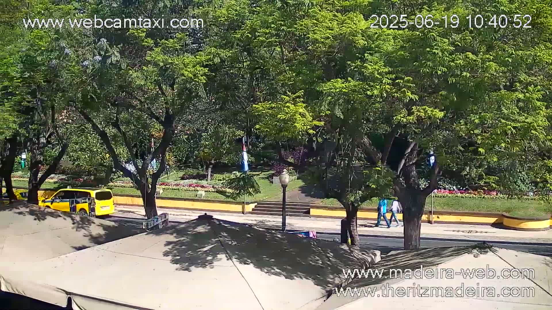 A sunny day reveals a yellow van parked near a park with lush green trees, flowers, and a paved walkway where two people stroll.