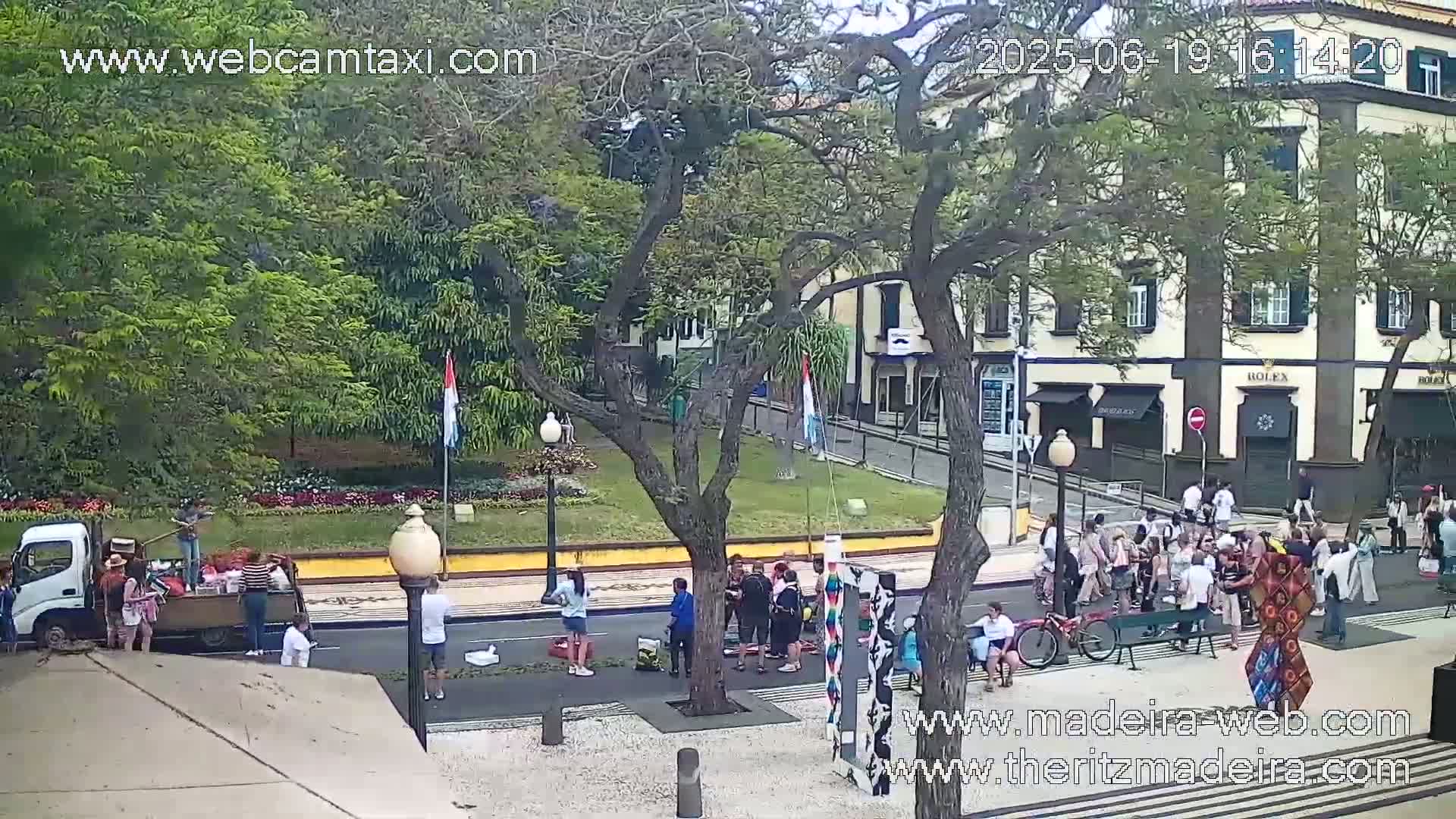 A sunny day shows a town square with people gathered, a truck being unloaded, and several buildings lining the street.